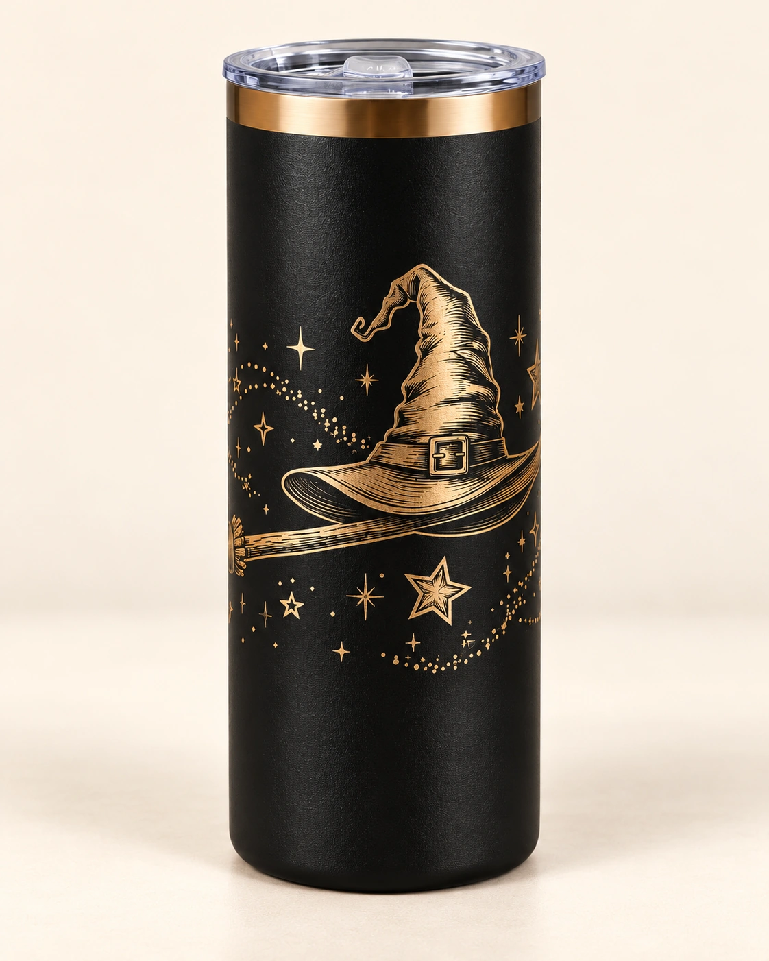 Termo negro con grabado dorado de sombrero de bruja, escoba y estrellas, sobre fondo marfil.